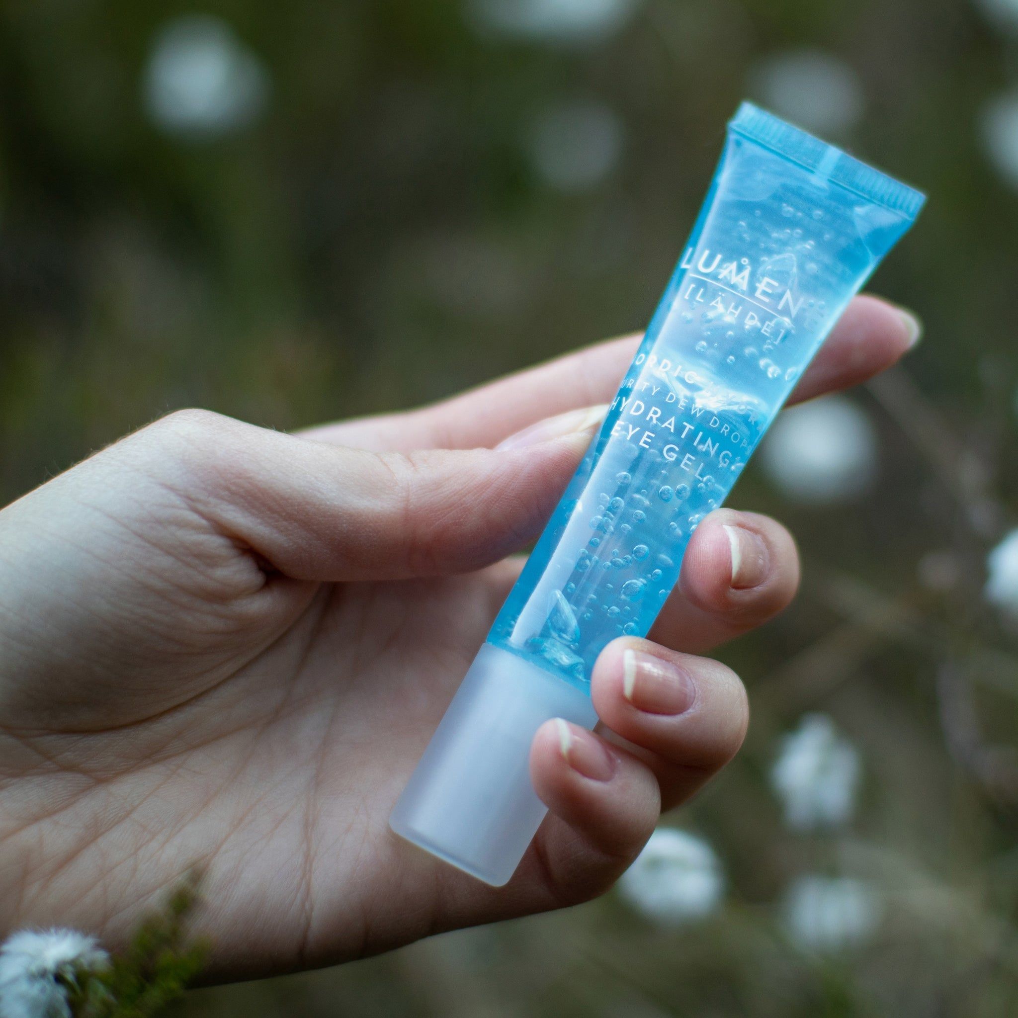 Hand hält eine blaue Tube. Aufschrift: Lumene [LAHDE] Nordic Hydra Purity Dew Drops Hydrating Eye Gel. Hintergrund: unscharfe Blüten.