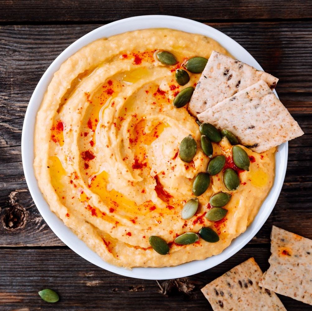 Hummus in weißer Schüssel. Mit Olivenöl, Kürbiskernen und Crackern. Auf Holzuntergrund.