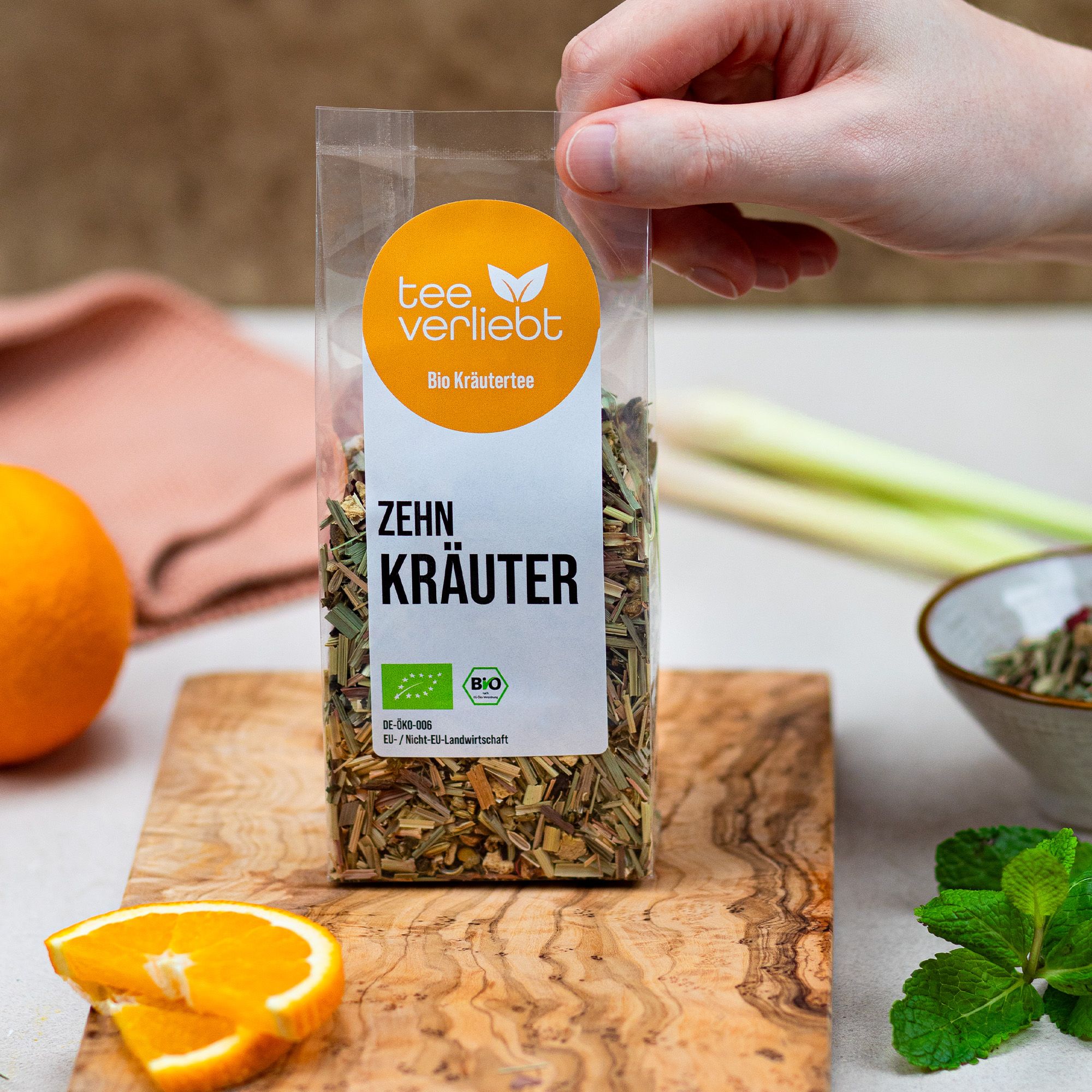 Hand hält Kräutermischung in transparenter Verpackung. Auf Holzbrett mit Orange, Minze und Zitrone. Logo und "Bio Kräutertee".