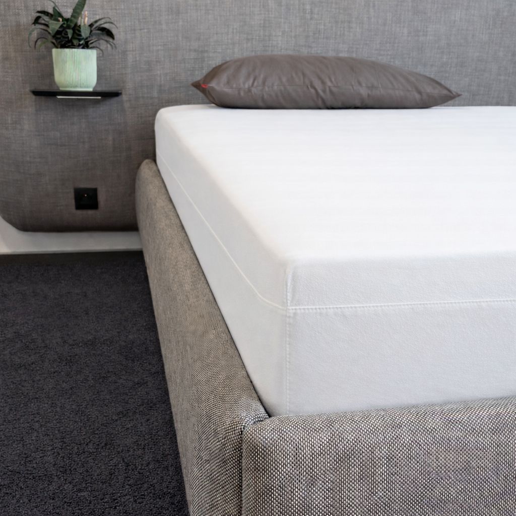 Weißes Spannbetttuch auf Bett. Graues Bettgestell, Kopfkissen. Zimmer mit Pflanze.