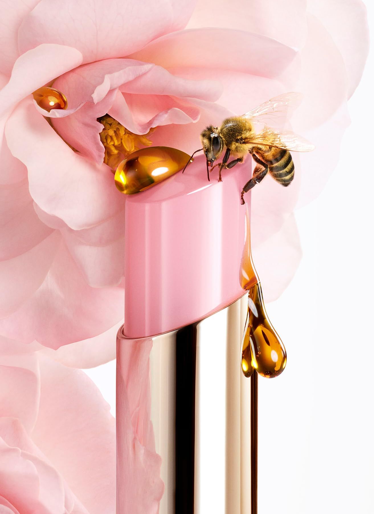 Ein Lippenbalsam-Stift mit rosa Hülle, goldfarbenen Details und Honigtropfen. Eine Biene sitzt auf dem Stift und berührt die Honigtropfen.