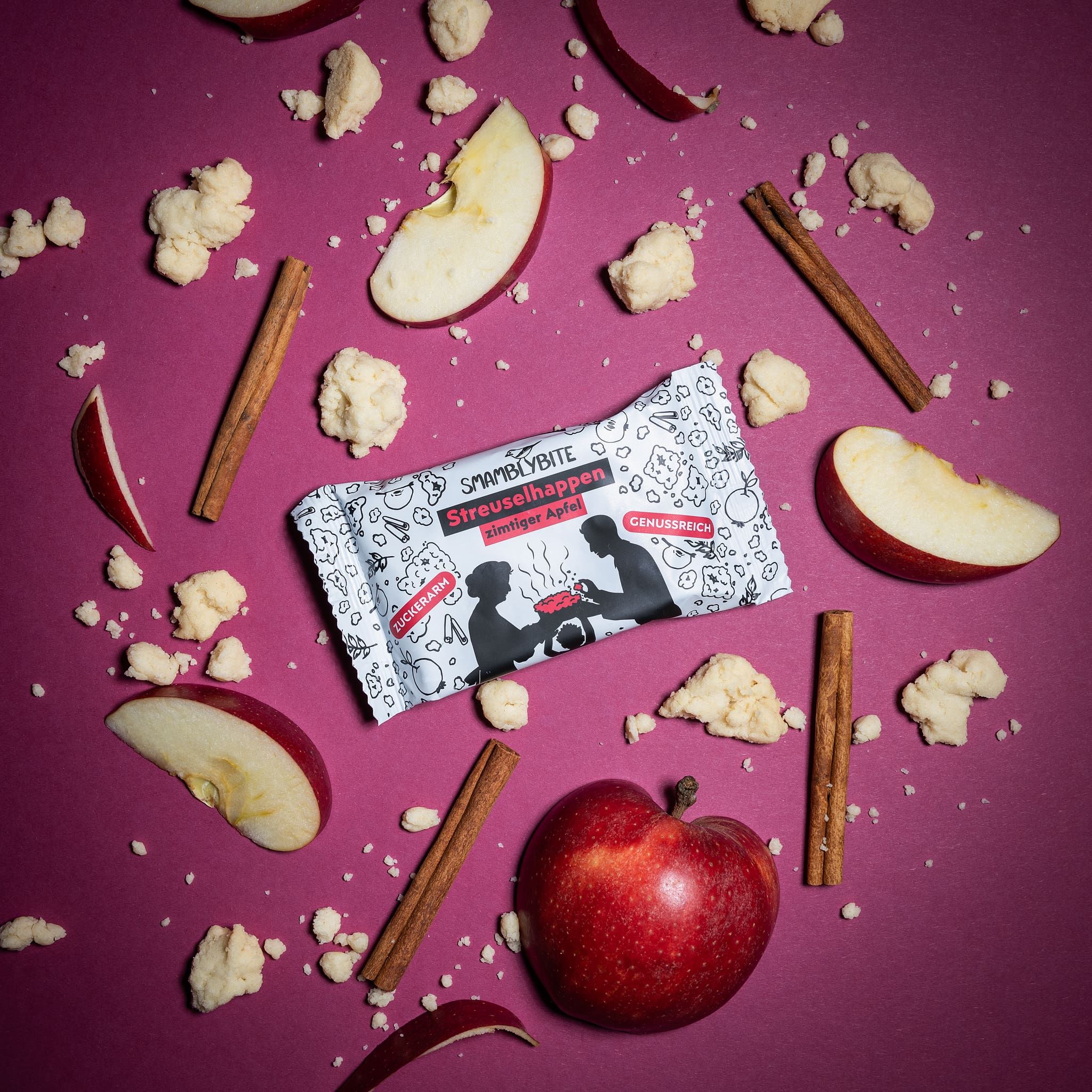 Streuselhappen-Riegel, Apfelscheiben, Zimtstangen und Streusel auf lila Untergrund. Verpackung mit Aufdruck: SMAMBLYBITE, Streuselhappen, zimtiger Apfel.