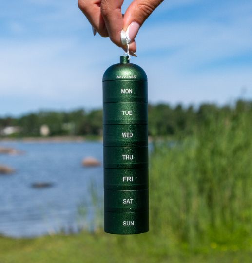 Grüne Pillendose mit 7 Fächern, gehalten vor einer natürlichen Kulisse mit Wasser und Gras.