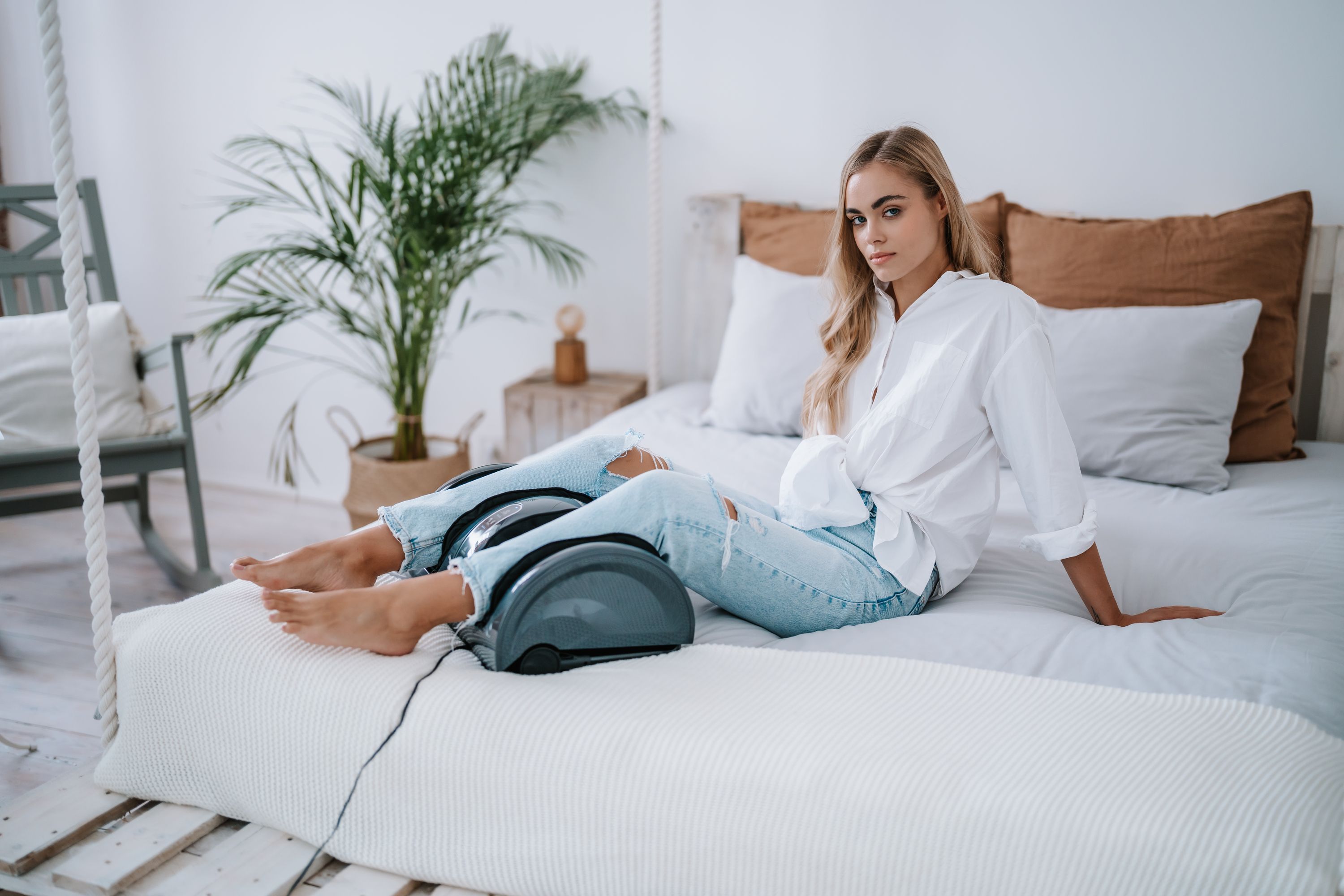 Eine Frau liegt auf einem Bett und benutzt ein Fußmassagegerät. Das Gerät ist dunkelgrau und hat zwei Öffnungen.