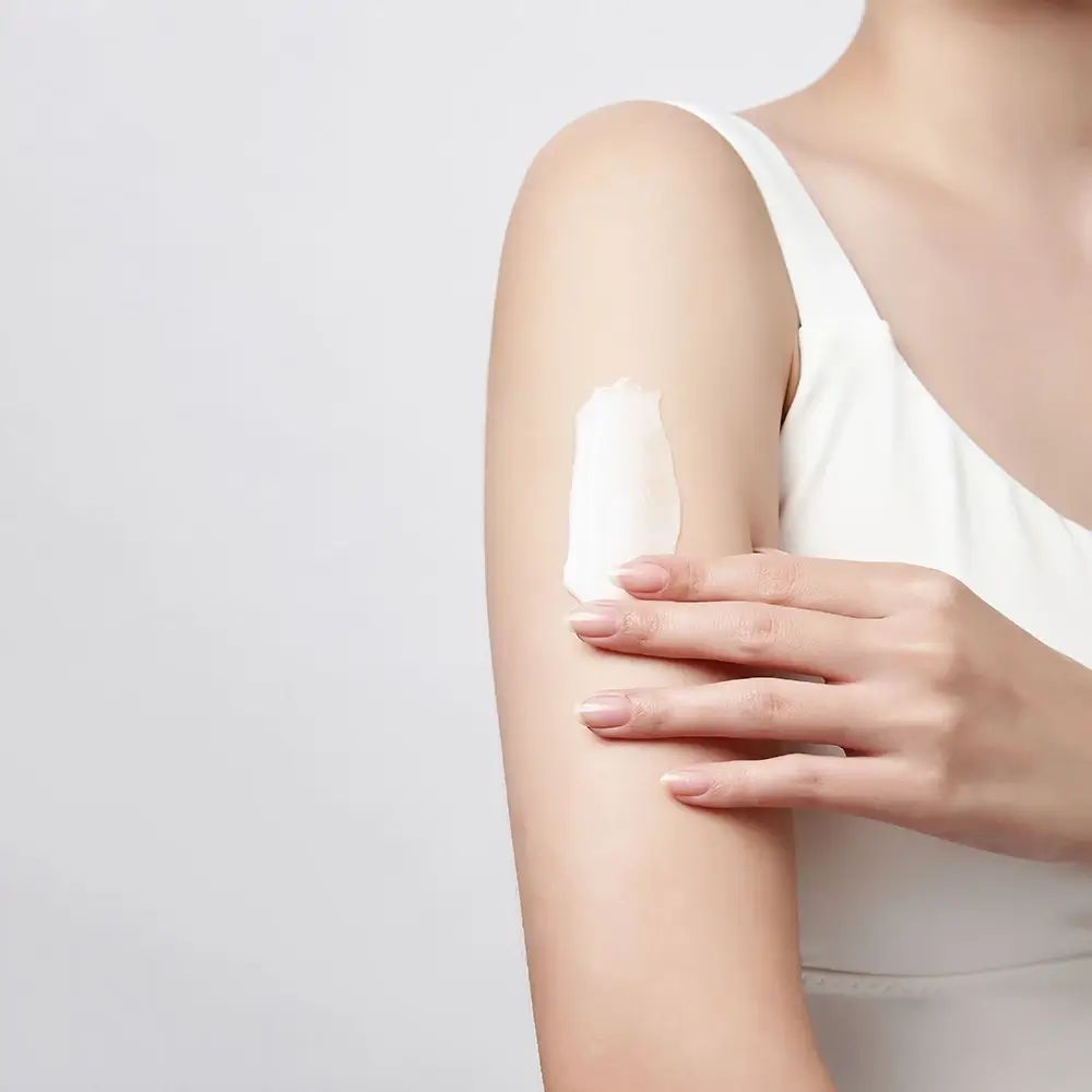 Arm mit weißer Creme. Die Creme wird auf die Haut aufgetragen. Person trägt ein weißes Tanktop.