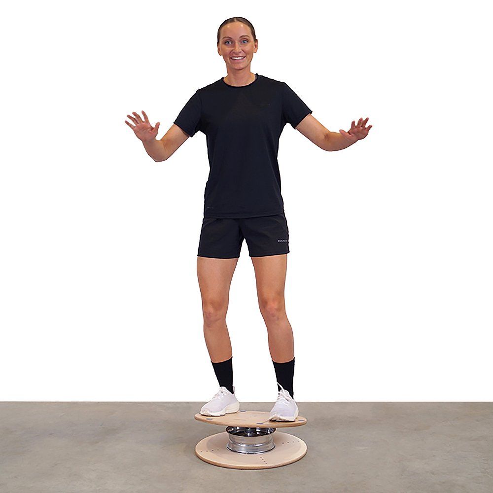 Person steht auf einem Pedalo Gleichgewichtstrainer. Das Gerät besteht aus Holz und Metall. Die Person balanciert mit ausgestreckten Armen.