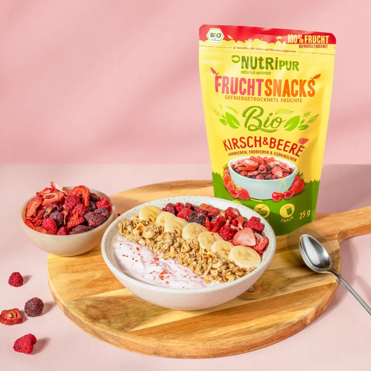 Beutel mit gefriergetrockneten Früchten, Kirschen & Beeren. Schale mit Müsli, Bananen und Früchten. Schale mit Früchten, Löffel.