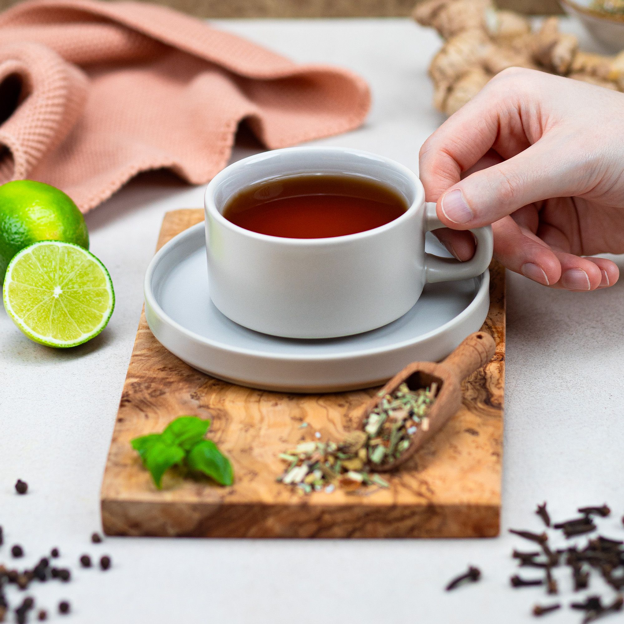 Tasse Tee auf Untertasse, Holzbrett, Limette, Kräutertee-Mischung, Holzlöffel. Hand hält Tasse.