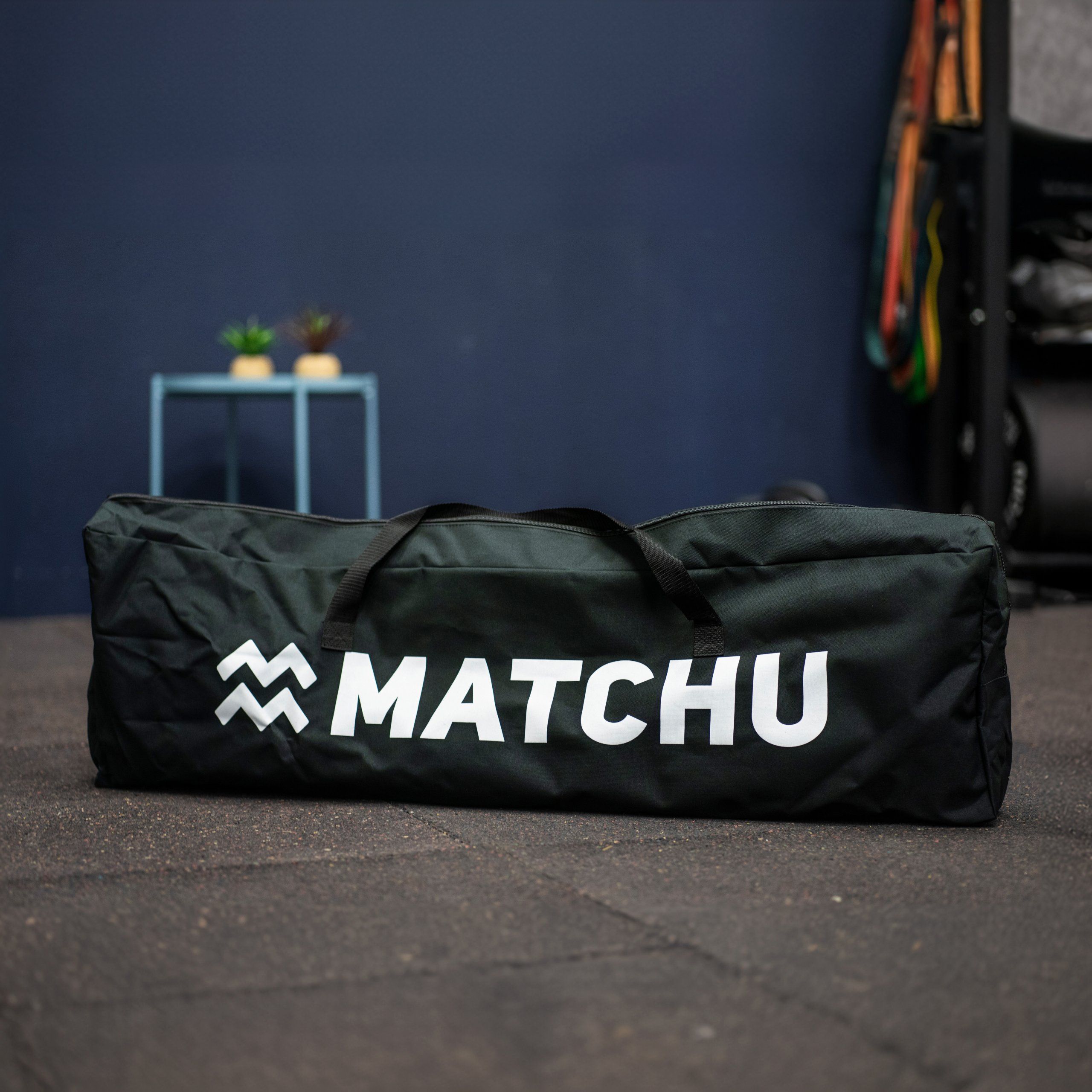 Schwarze Tragetasche mit weißem Logo. Marke: MATCHU. Steht auf dem Boden in einem Fitnessstudio.