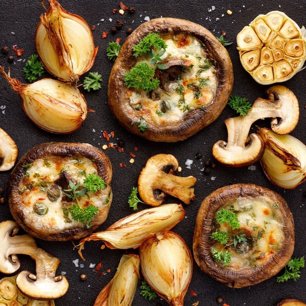 Gefüllte Pilze, Zwiebeln und Knoblauch auf schwarzem Untergrund. Kräuter als Garnierung.