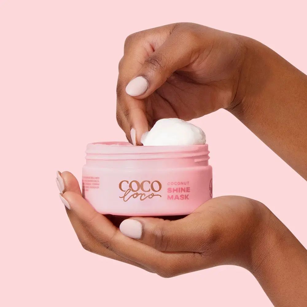 Hände halten einen geöffneten, rosa Coco Loco-Tiegel. Creme wird entnommen. Aufschrift Coco Loco, Coconut Shine Mask.