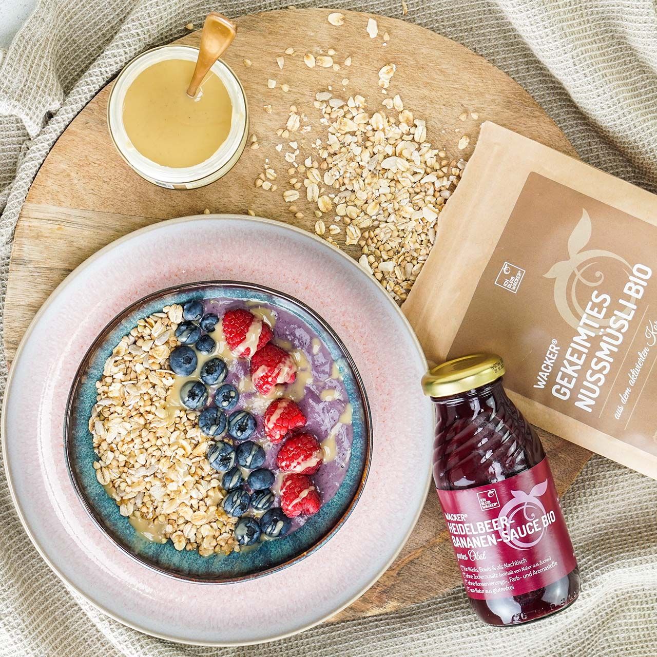 Schüssel mit Müsli, Beeren und Sauce. Flasche Heidelbeer-Bananen-Sauce Bio. Beutel Gekeimtes Nussmüsli Bio. Aufschrift: Wacker.