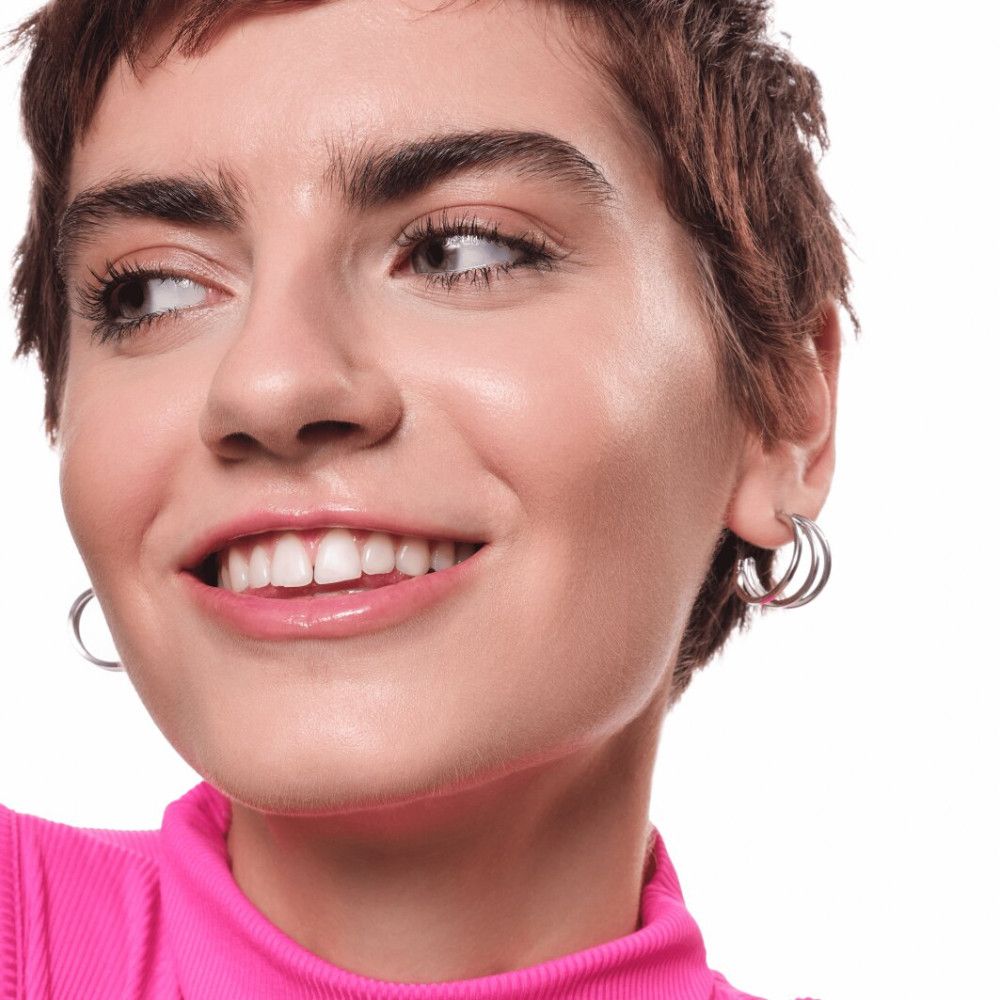 Porträt einer Frau mit kurzen Haaren, die in die Kamera lächelt. Sie trägt Ohrringe und ein pinkes Oberteil.