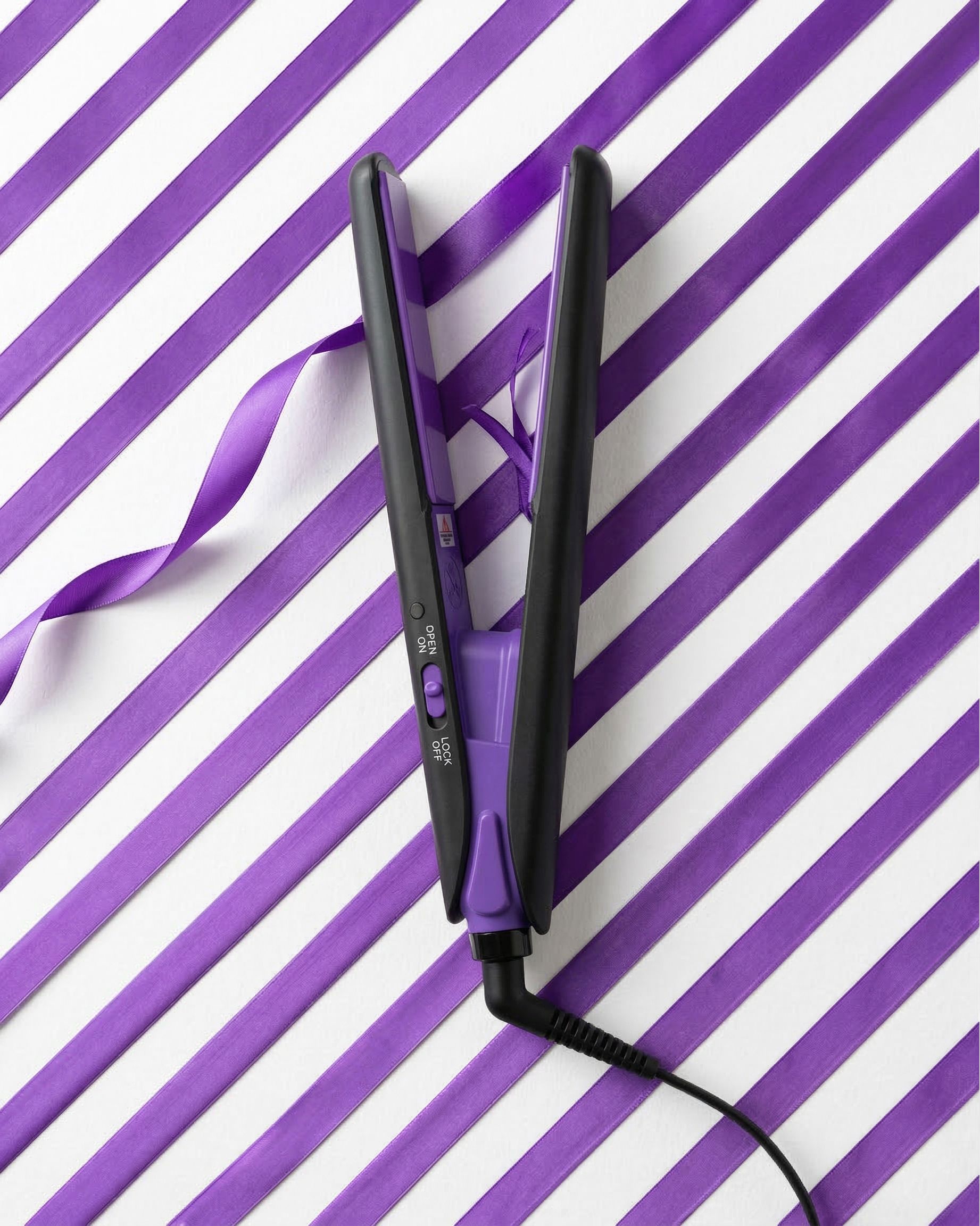 Schwarzer und violetter Mini-Haarglätter. Offene Platten, Kabelanschluss. Auf violett-weißem Streifenmuster.