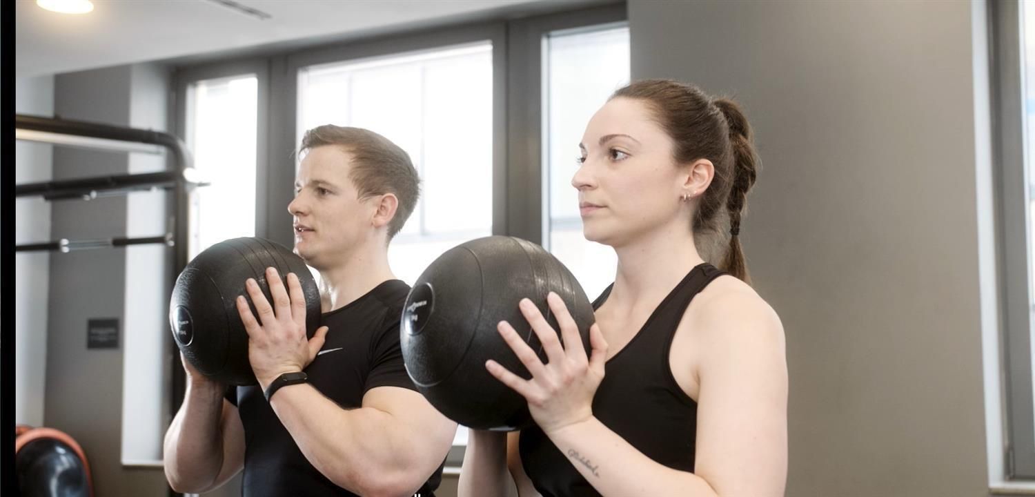 Zwei Personen halten jeweils einen schwarzen Slamball. Sie befinden sich in einem Fitnessstudio.