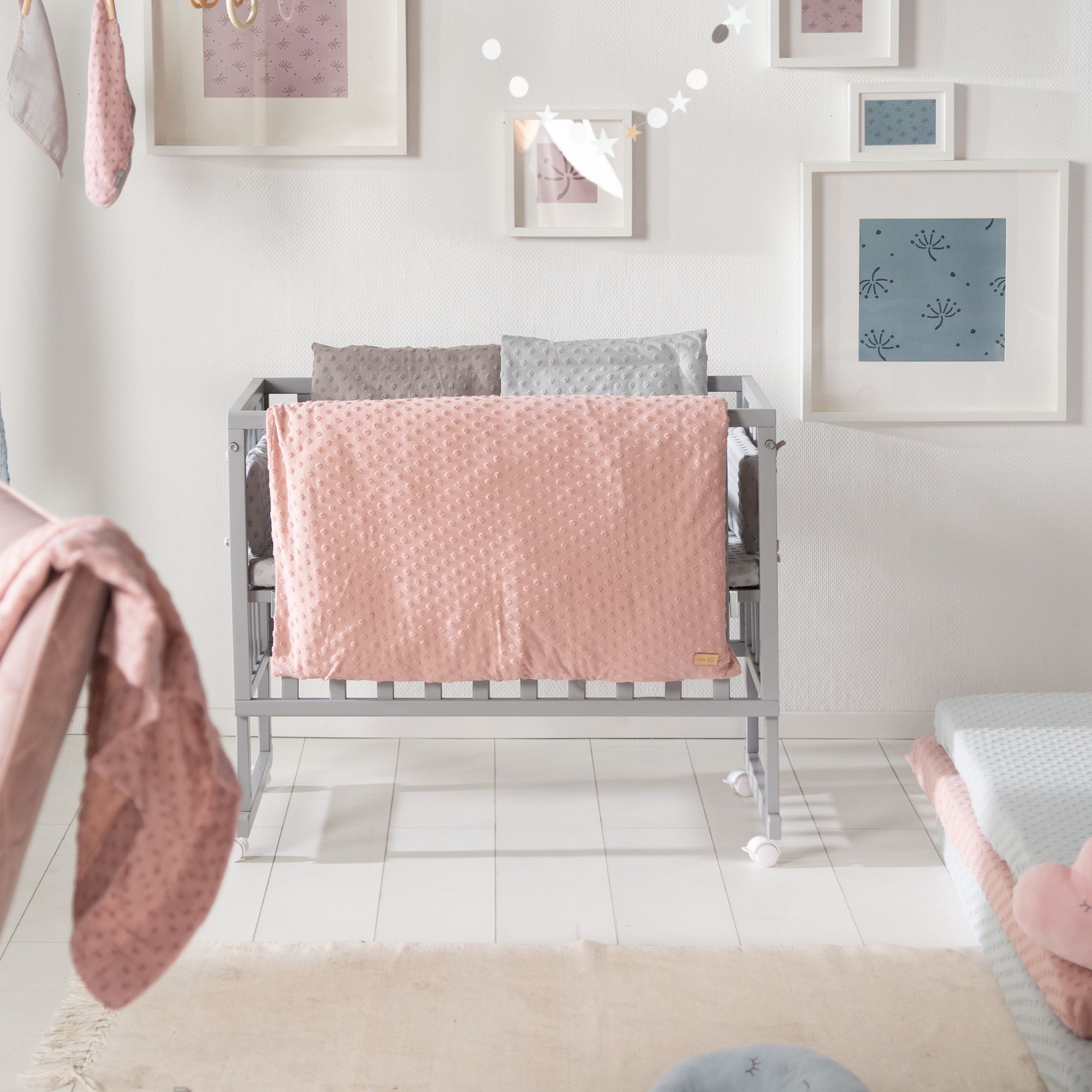 Babybettwäsche in Rosa, auf einem grauen Babybett. Im Hintergrund Wanddekoration.