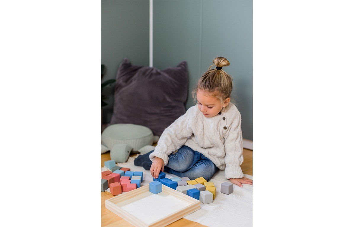 Kleines Mädchen spielt mit Holzbausteinen auf dem Boden. Bauklötze in einem Holzrahmen und lose daneben.