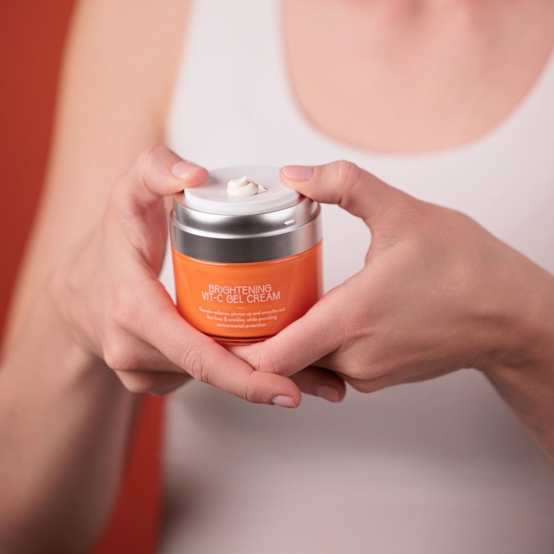 Hände halten eine orangefarbene Creme in einem Glas. Aufschrift: Brightening Vit-C Gel Cream. Text: Reveals radiance, plumps up and smooths out fine lines & wrinkles.