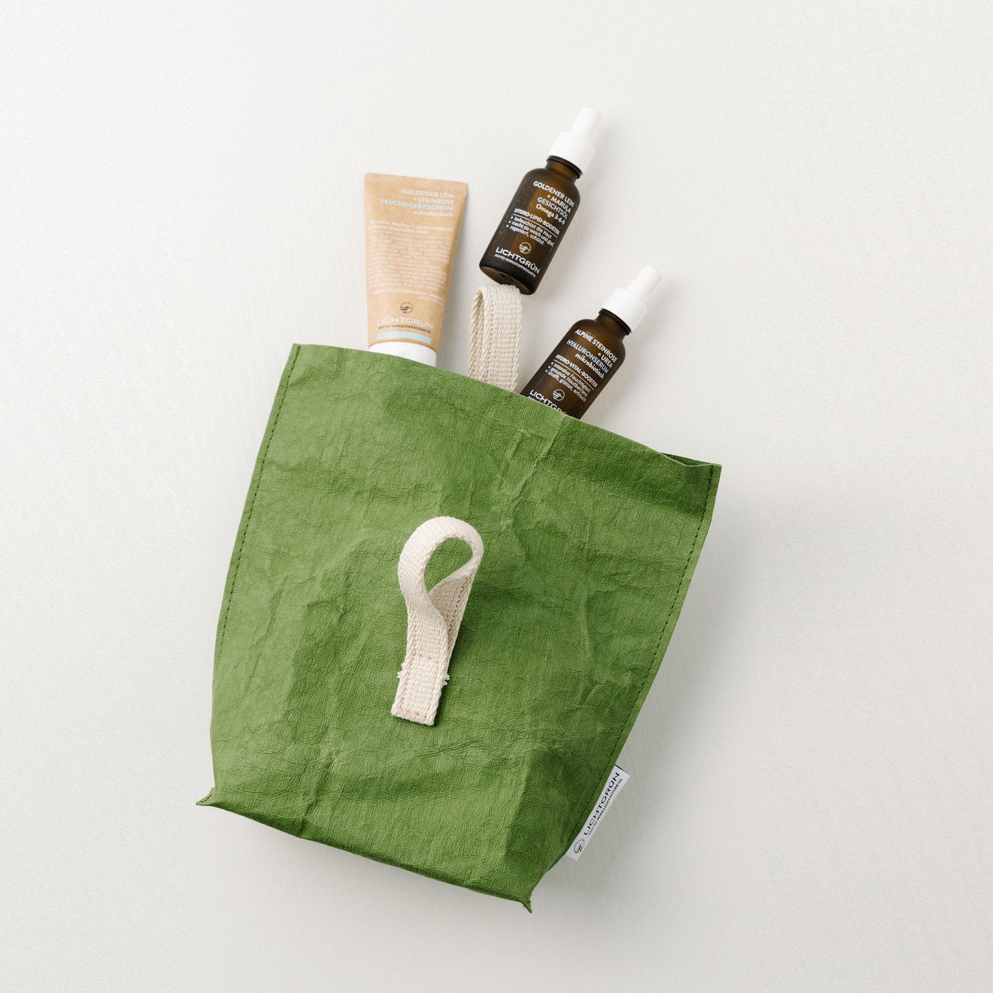 Grüne Tasche mit Griff, enthält zwei Flaschen und eine Tube. Produkte für die Hautpflege, auf hellem Hintergrund.