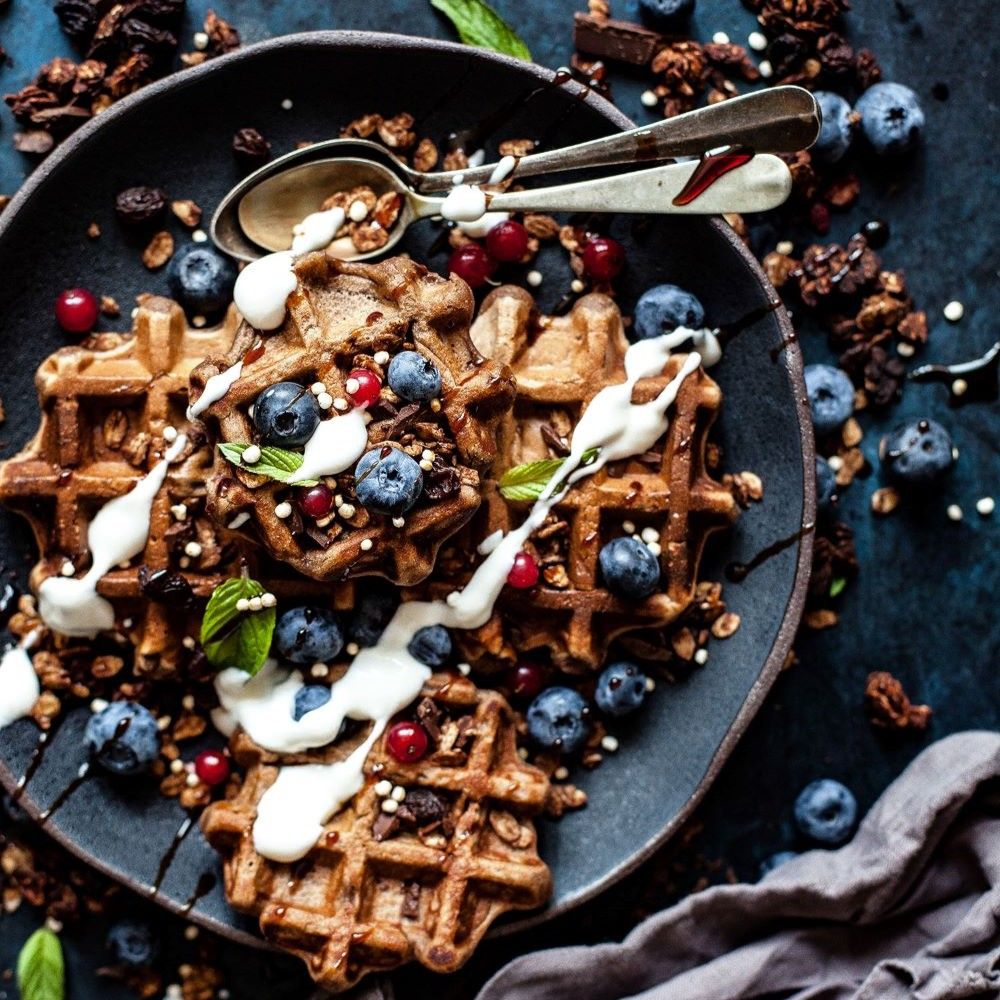 Waffeln auf einem Teller mit Blaubeeren, Granola und Creme. Zwei Löffel liegen auf dem Teller. Dunkler Hintergrund.