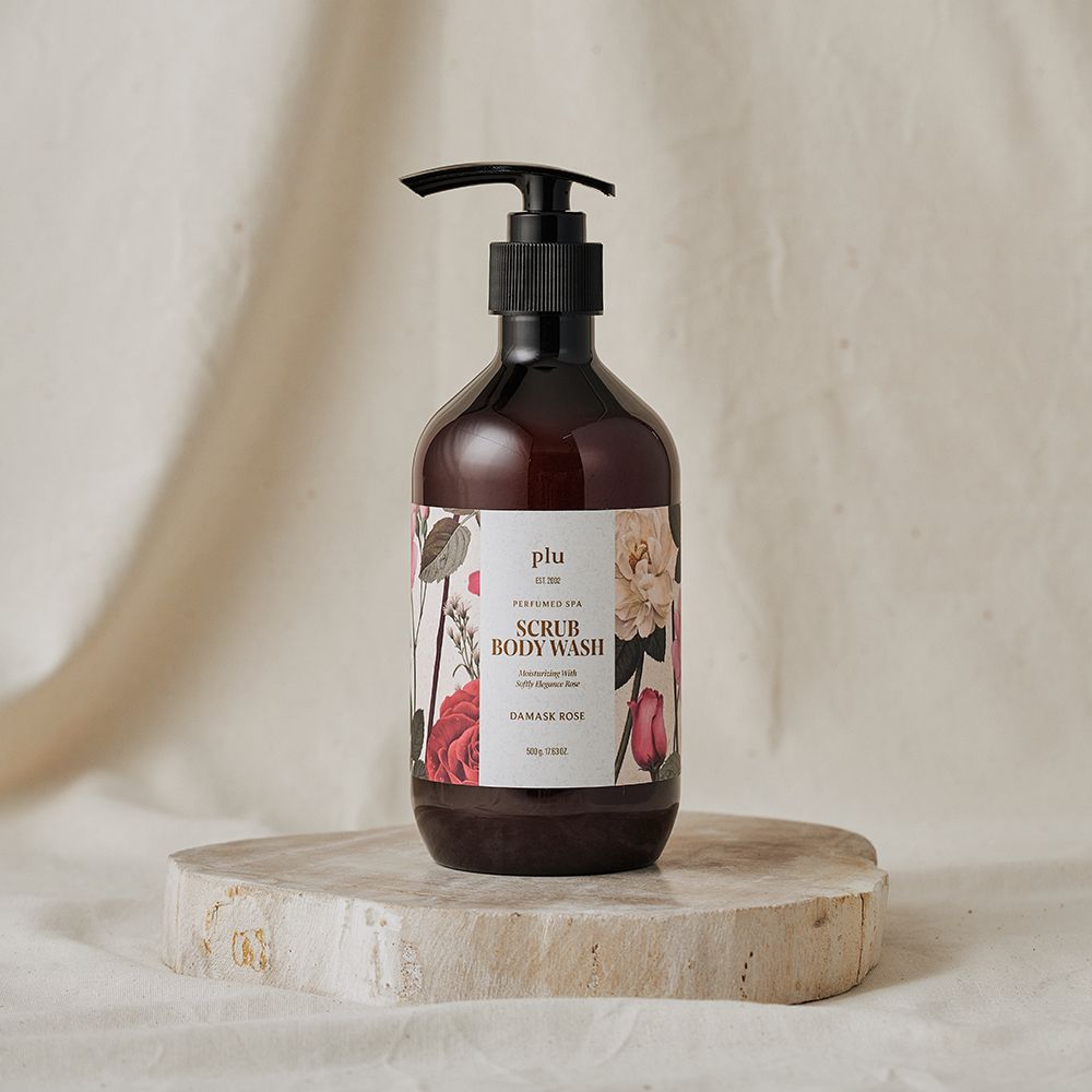 Braune Flasche mit schwarzem Pumpverschluss auf einem Stein. Etikett mit floralem Design und Schriftzug: Scrub Body Wash, Damask Rose. Marke: plu.