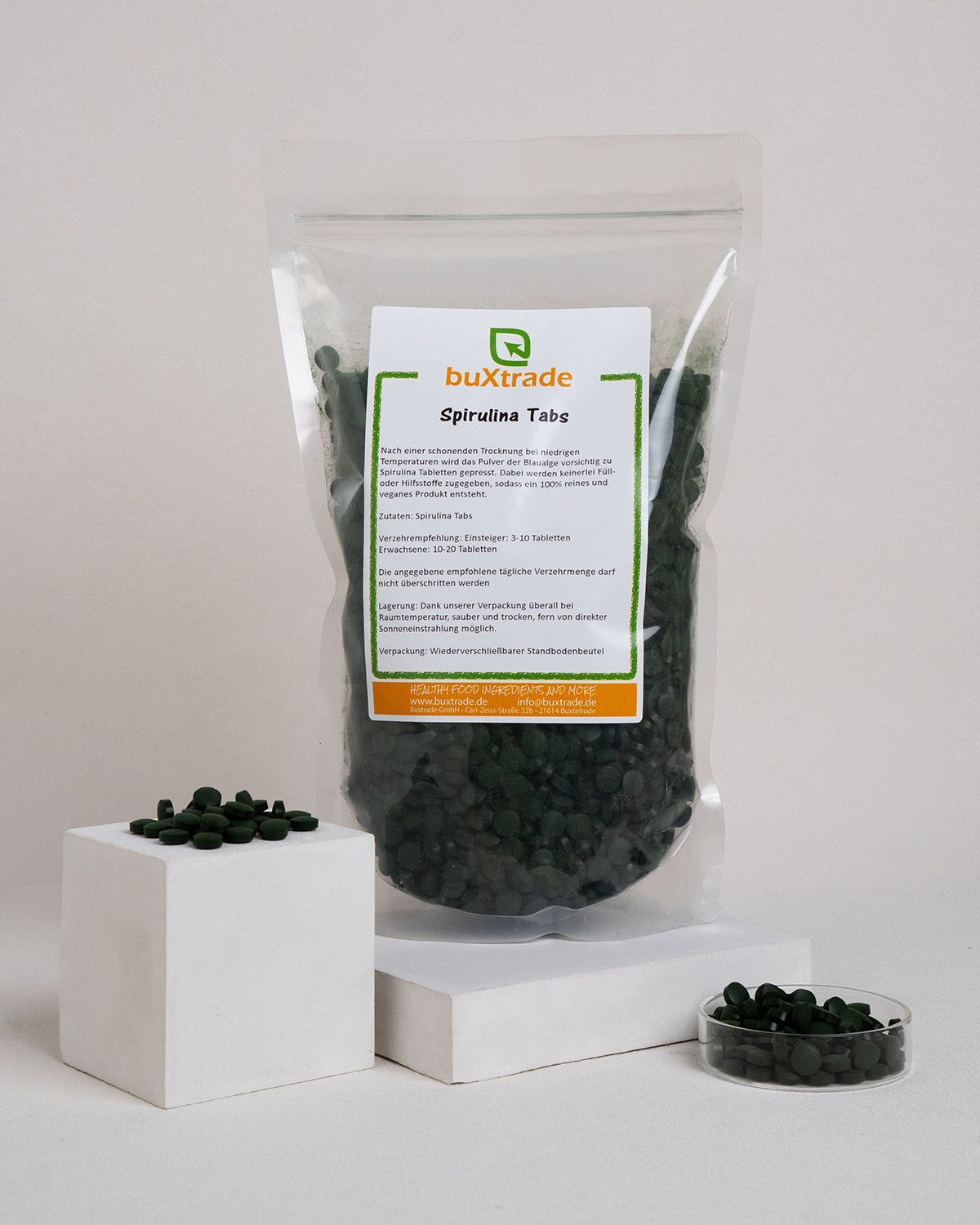 Transparente Tüte mit grünen Tabletten, Etikett und Glasschale. Etikett mit Text: buXtrade Spirulina Tabs.