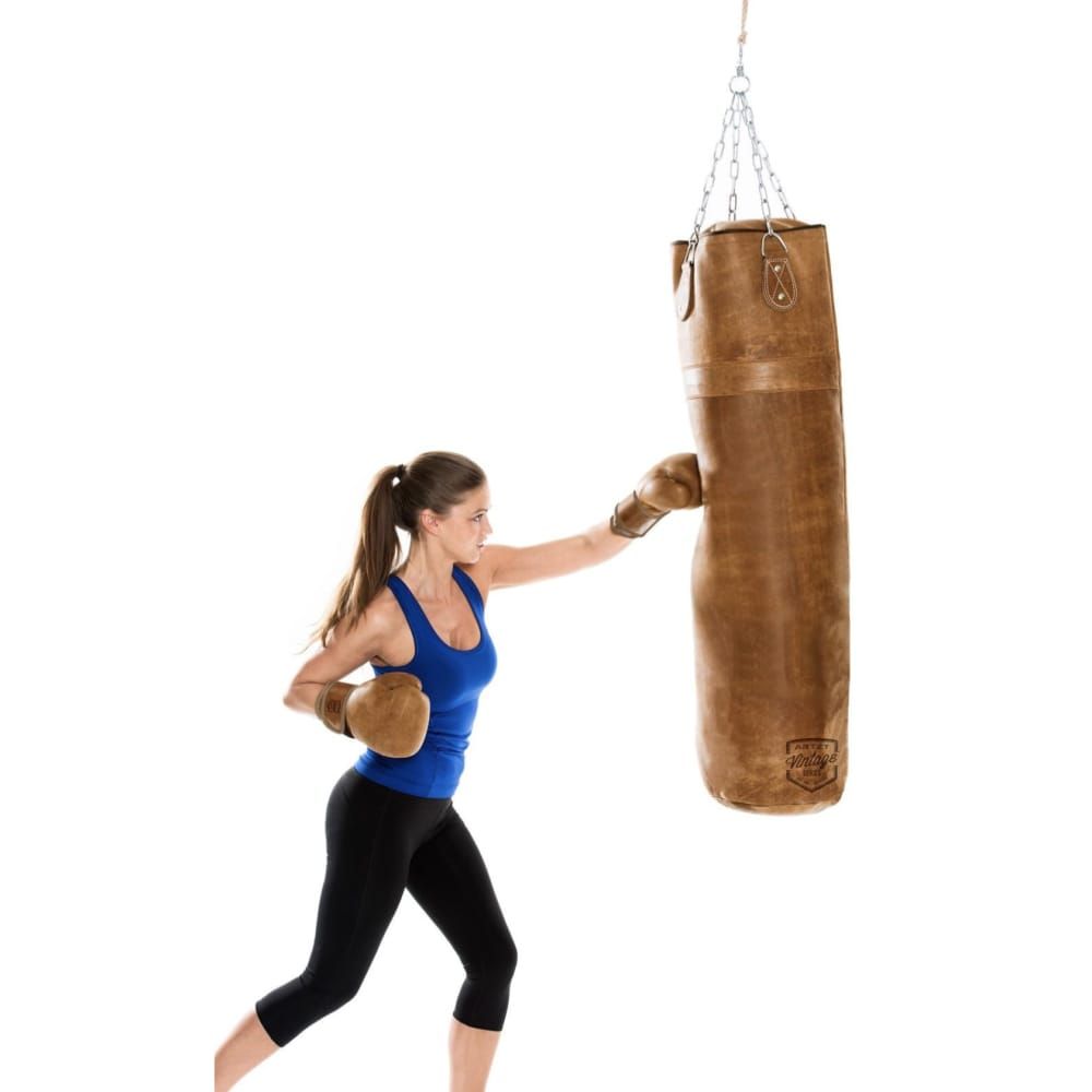 Brauner Leder-Boxsack, von Frau geschlagen. Vintage-Look. Frau trägt Boxhandschuhe und Sportkleidung.