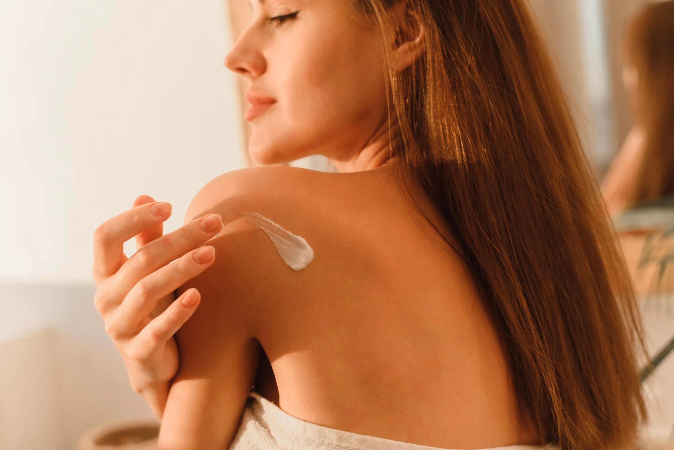 Eine Frau trägt Creme auf ihre Schulter auf. Helle Haut, braune Haare, Sonnenlicht.