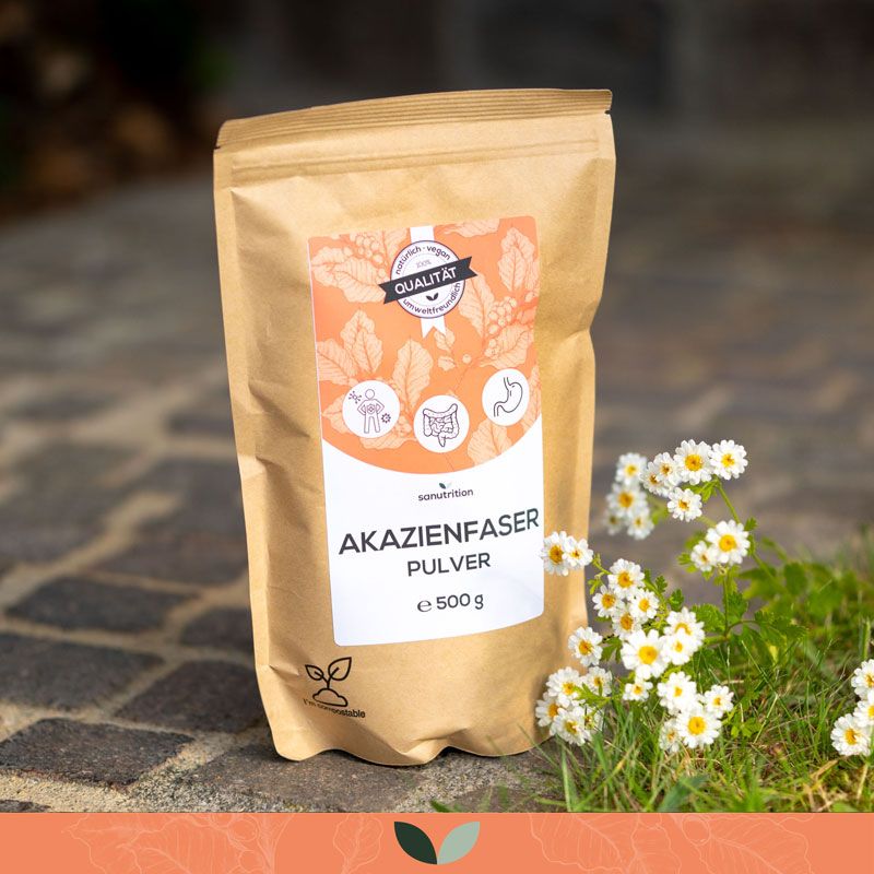 Beutel Sanutrition Akazienfaser Pulver, 500g, auf Pflastersteinen. Blüten im Hintergrund. Qualitätssiegel.