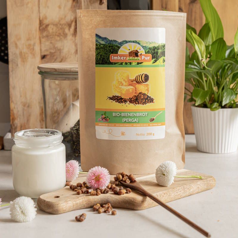 Auf einem Holzbrett: Bienenbrot, Milch, Honig, Blumen. Im Hintergrund: Verpackung mit Aufkleber: ImkerPur, Bio-Bienenbrot (Perga).