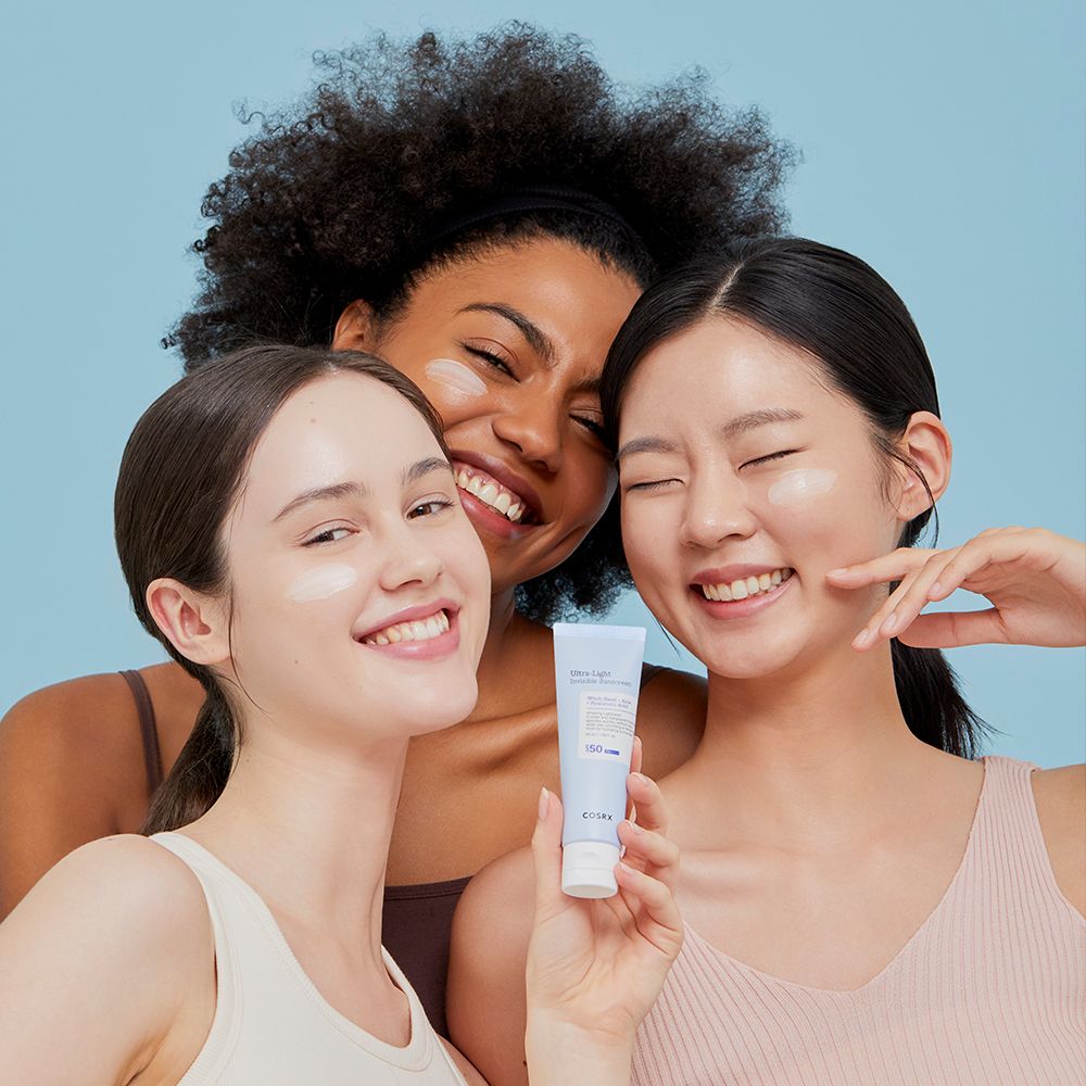 Drei Frauen lächeln und halten die COSRX Ultra Light Invisible Sunscreen Tube.
