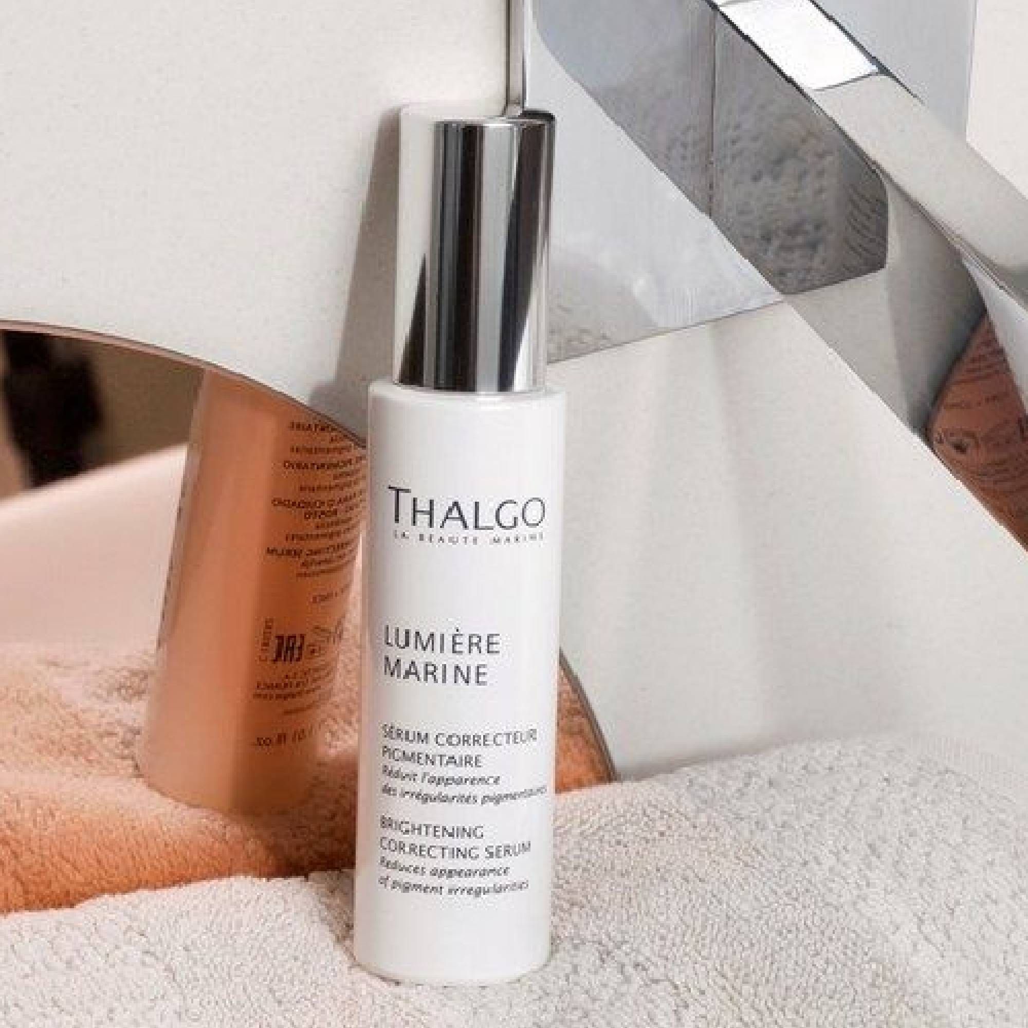 Weißes Serum mit silbernem Verschluss. Text: THALGO, LUMIÈRE MARINE, Serum correcteur pigmentaire, BRIGHTENING CORRECTING SERUM. Auf Handtuch.