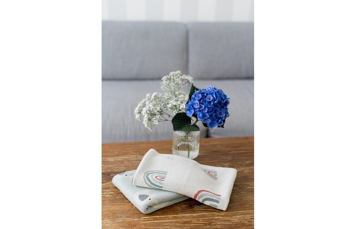 Zwei gefaltete Decken mit Regenbogenmuster auf einem Tisch. Beige mit bunten Regenbogenmotiven. Auf dem Tisch stehen Blumen.