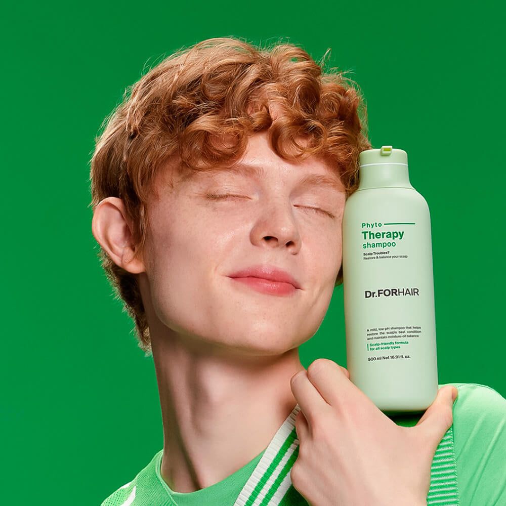 Mann mit roten Locken hält eine Flasche Dr. FORHAIR Phyto Therapy Shampoo vor einem grünen Hintergrund.