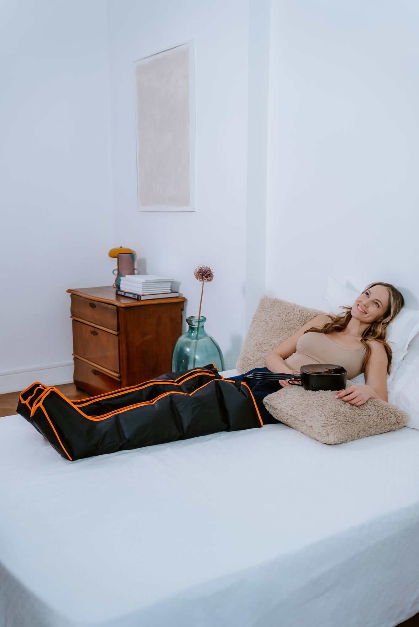 Frau liegt auf dem Bett mit Bein-Lymphmassagegerät. Schwarzes Gerät mit orangefarbenen Akzenten. Steuerung daneben.