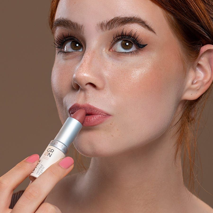 Frau hält einen rosafarbenen Lippenstift. Silberfarbener Schaft, rosafarbener Stift. Aufschrift: GRN, Lipstick, certified organic. Frau trägt rosa Nagellack.