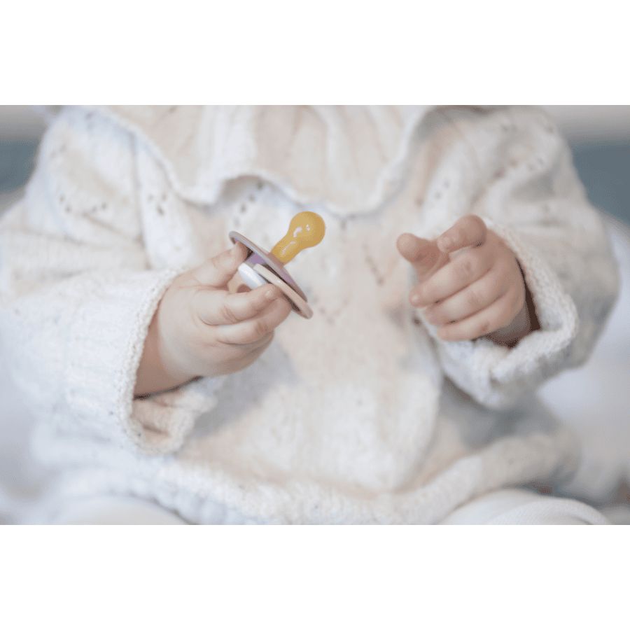 Baby hält Schnuller. Gelber Sauger, weißer Schild. Baby trägt weißen Pullover.