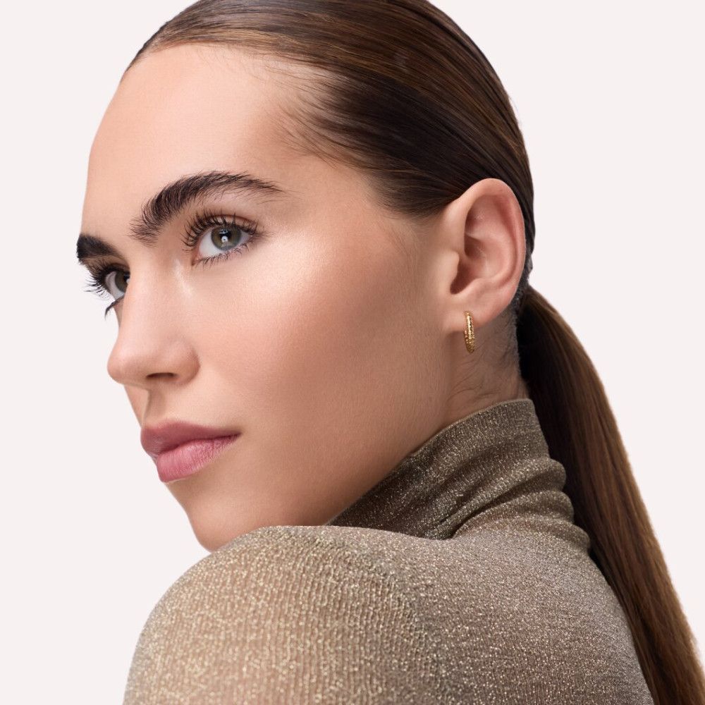 Porträt einer Frau mit goldfarbenem Oberteil. Sie trägt goldene Creolen und hat einen Pferdeschwanz.