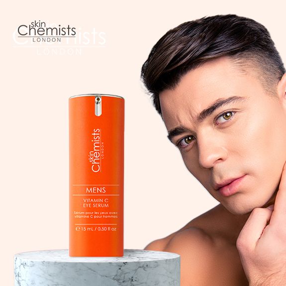 Mann mit Serumfläschchen. Aufschrift: skinChemists LONDON, MENS Vitamin C EYE SERUM, 15 ml / 0.50 fl oz. Fläschchen auf Podest.