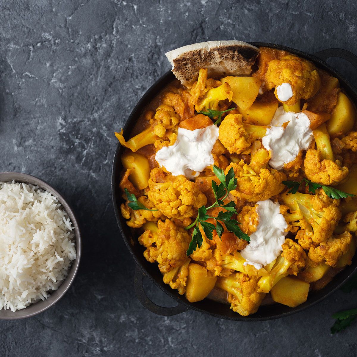 Gericht mit Blumenkohl, Kartoffeln und Soße in einer schwarzen Pfanne. Eine Schüssel mit Reis steht daneben.