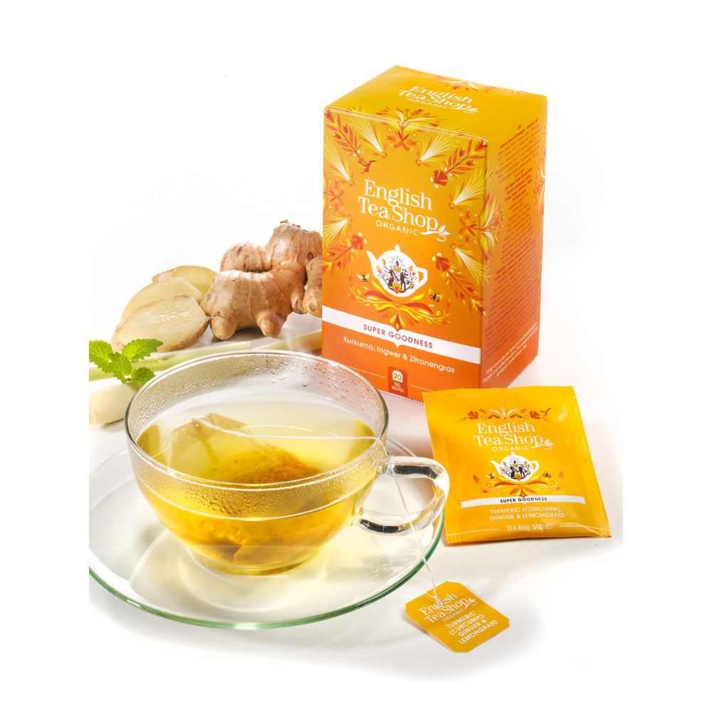 Glas-Teetasse mit Teebeutel und Aufguss. Daneben Teebox und Teebeutel-Verpackung. Ingwer und Minze als Dekoration.