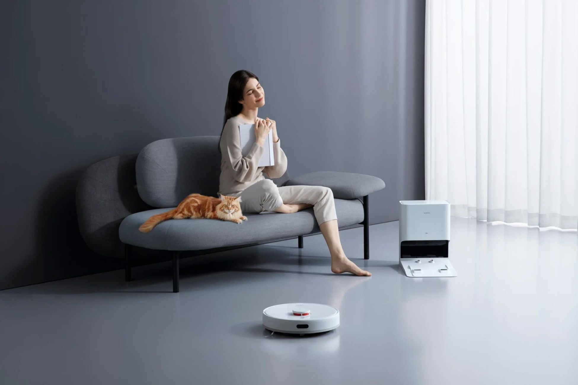 Frau sitzt auf einem Sofa mit Katze und Roboterstaubsauger. Die Ladestation ist im Hintergrund zu sehen.