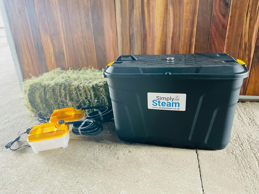 Schwarzer Behälter mit gelben Griffen, Simply Steam-Logo. Zwei gelb-weiße Geräte mit Kabeln. Heuballen daneben. Holzverkleidung im Hintergrund.