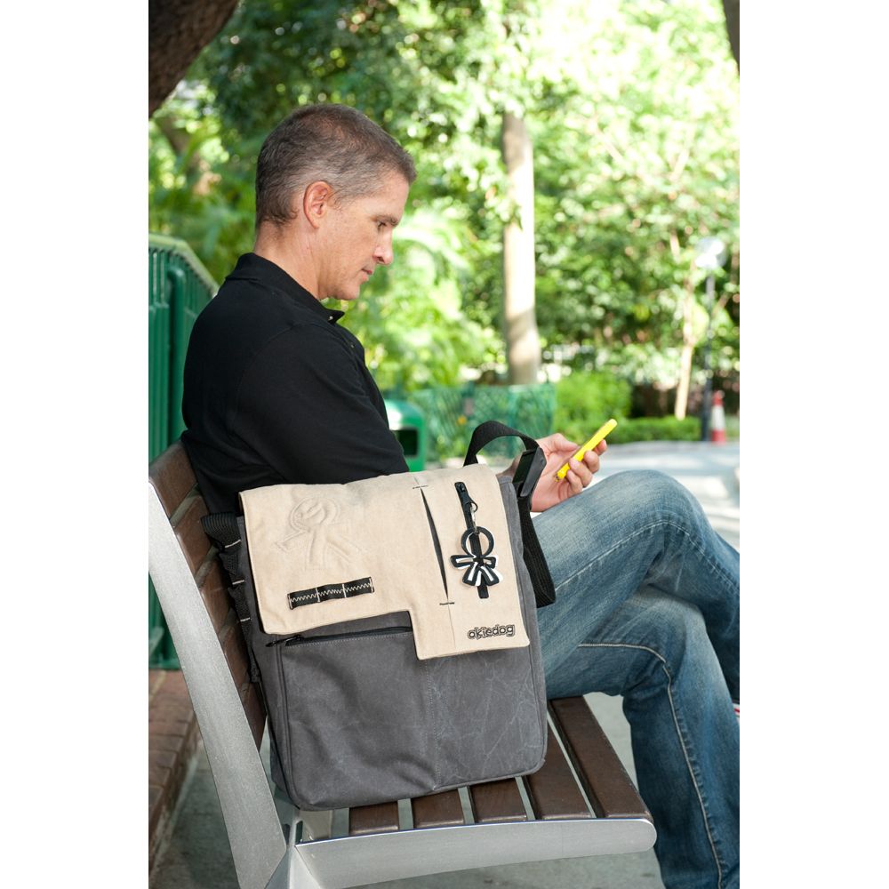 Mann sitzt mit Okiedog Wickeltasche Loft Paige auf Bank. Tasche beige-grau, Logo und Anhänger sichtbar.