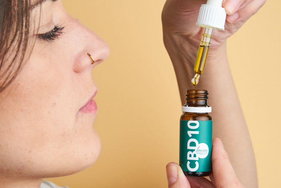 Person hält eine Flasche mit grüner Etikettierung und Pipette. Aufschrift CBD10. Tropfen fallen in den Mund.
