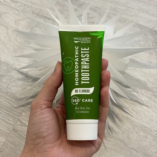 Grüne Zahnpasta-Tube in Hand gehalten. Aufschrift: Homeopathic Toothpaste, No Fluoride, 360° Care. Logo: Zahn. Hintergrund: weiße Dekoration.