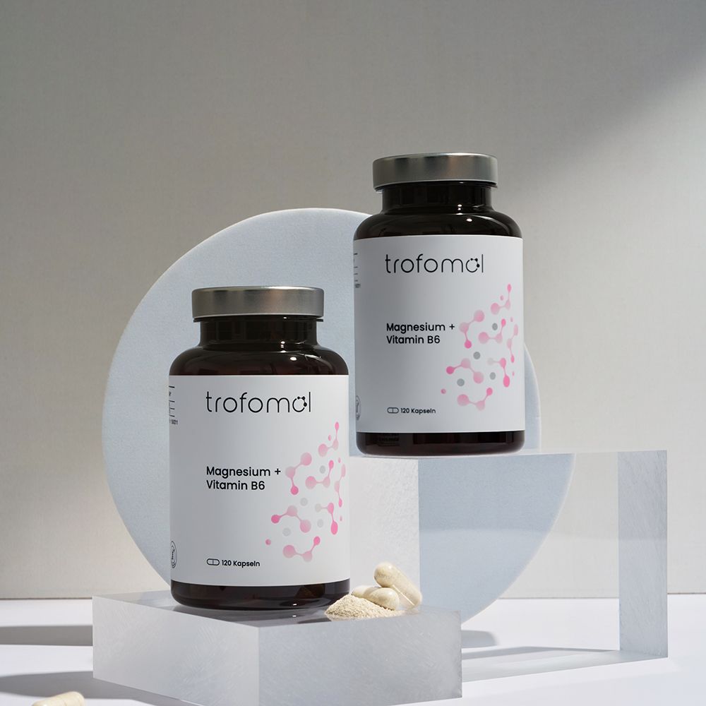 Zwei braune Glasflaschen mit weißem Etikett. Aufschrift: trofomol, Magnesium + Vitamin B6. Kapseln liegen daneben.
