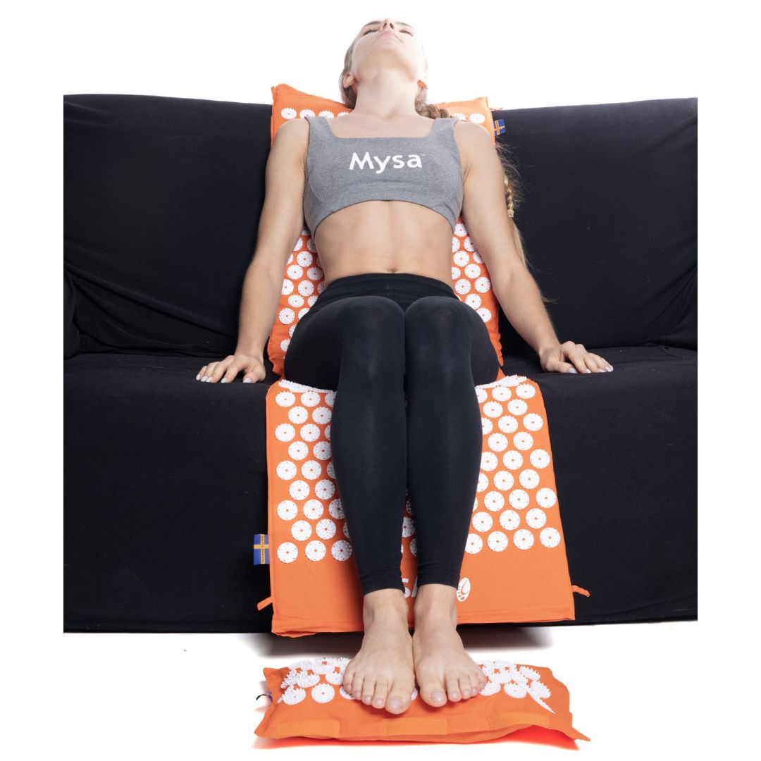 Frau sitzt auf einer schwarzen Couch, auf einer orangefarbenen Akupressurmatte und einem Kissen. Das Produkt ist sichtbar.