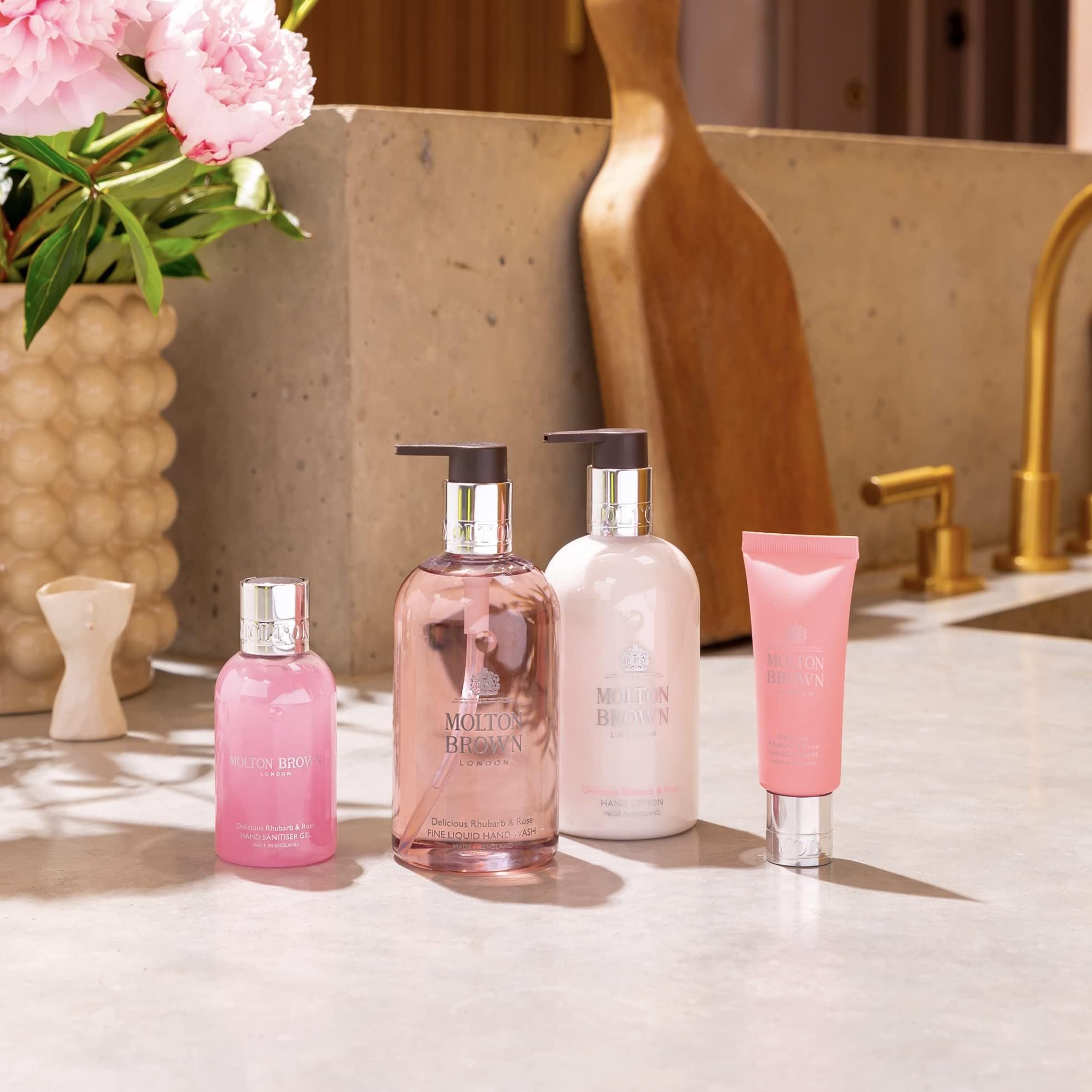 Rosa Produkte auf einer Theke: Handcreme, Seife, und Parfüm. Blumenarrangements im Hintergrund. Goldfarbene Wasserhähne.