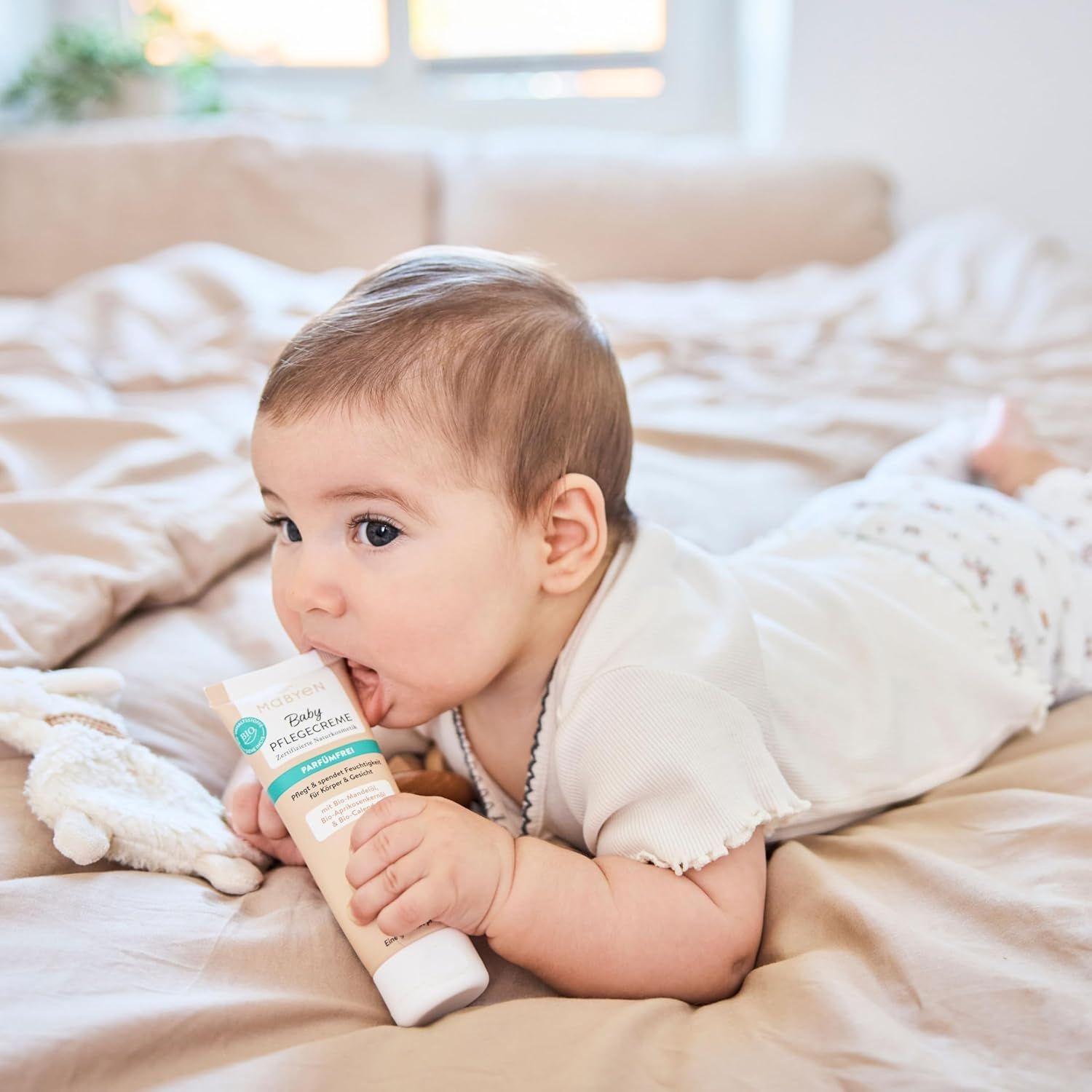 Ein Baby liegt auf dem Bauch und beißt auf eine Tube MABYEN Baby Pflegecreme. Die Tube ist beige mit grünen und weißen Akzenten.